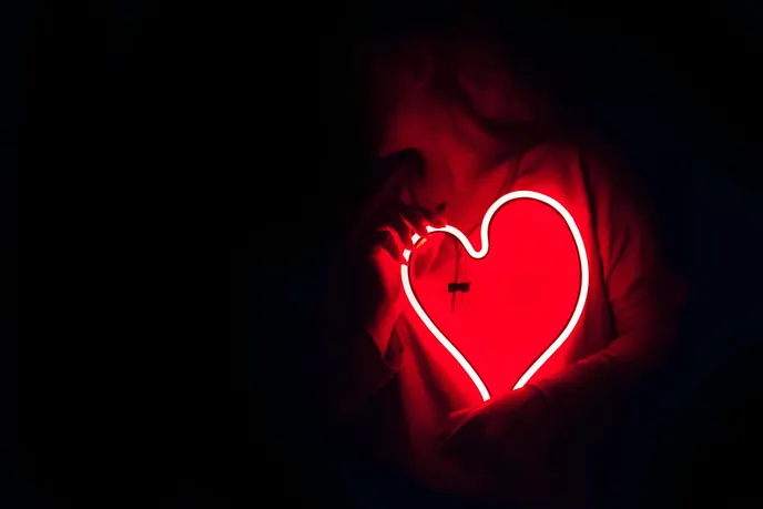 A person holding a glowing red neon heart, symbolizing love and affection, set against a dark background.