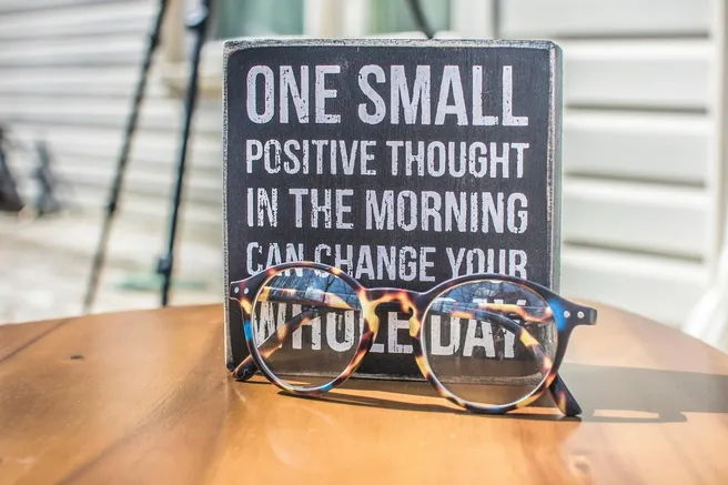 A decorative sign that reads 'One small positive thought in the morning can change your whole day' placed next to a pair of stylish eyeglasses on a wooden table. The background shows blurred outdoor elements, suggesting a bright, peaceful setting for reflection and motivation.